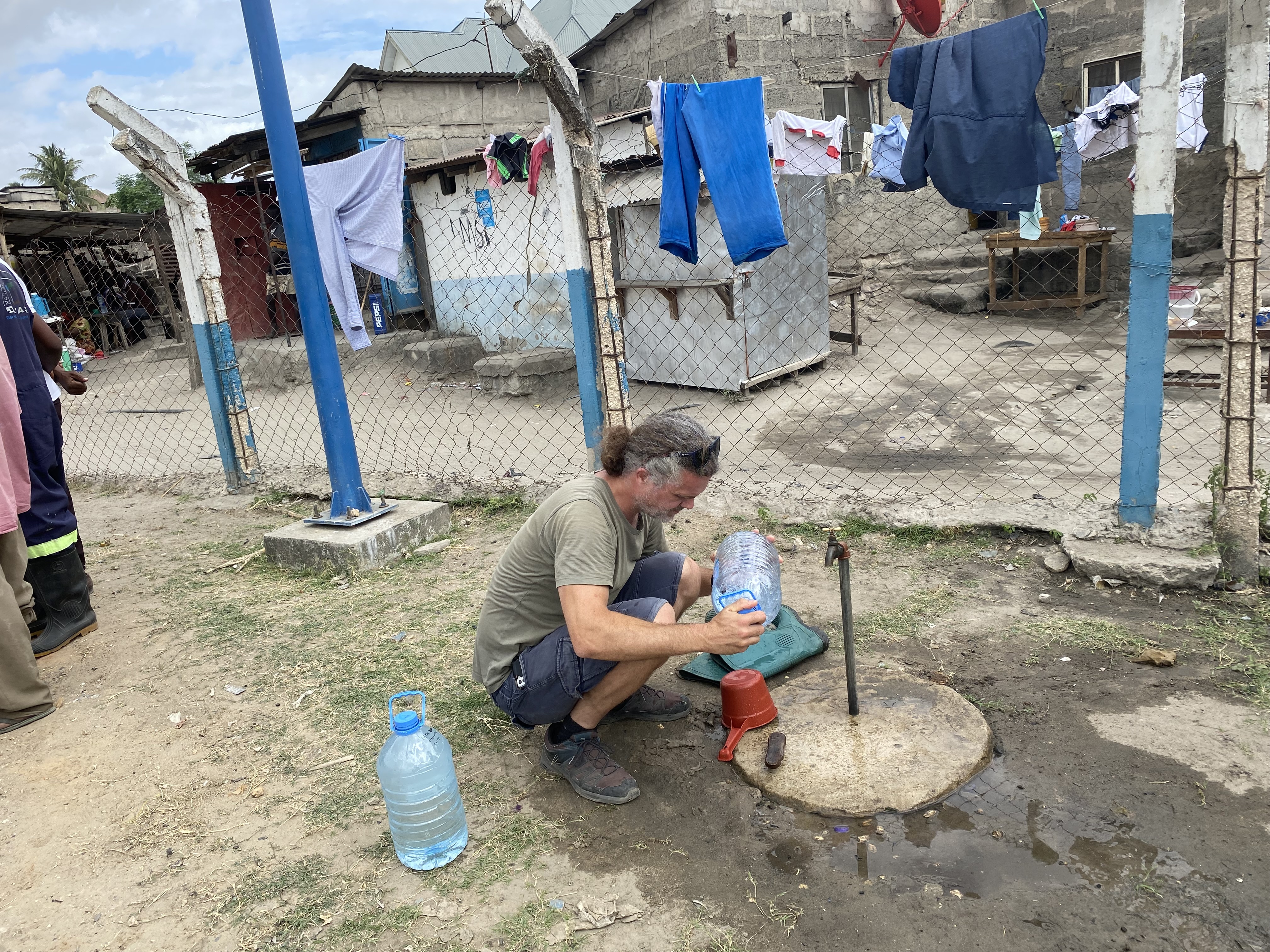 , Sampling of drinking water in Dar es Salaam in very close proximity to an open wastewater treatment plant. (Photo: Adriana Krolicka), IMG 8174, <p>Adriana Krolicka</p>, A man holding a water container, sitting on his knees in front of a water post.