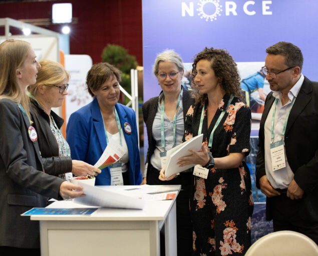 Thomas Hovmøller Ris, Forskningsleder for gruppen Fiskebiologi og akvakultur Naouel Gharbi presenterer på NORCE sin stand., Stand1, <p>Thomas Hovmøller Ris, NORCE</p>, Seks personer står rundt et lite bord og diskuteter et papir.